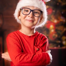 Pan Aparacik Dawid Kubica - Uśmiechnięty chłopiec w okularach i czerwonym swetrze, ubrany w czapkę Świętego Mikołaja z puszystym pomponem, pozuje na tle rozświetlonej choinki.