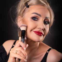 Pan Aparacik Dawid Kubica - Uśmiechnięta blondynka z wyrazistym makijażem oczu i czerwonymi ustami trzymająca pędzle do makijażu przy policzku, na czarnym tle.