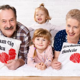 Pan Aparacik Dawid Kubica - Uśmiechnięta rodzina: dziadek w koszuli w kratę z poduszką 'Kocham Cię Dziadku', babcia mrugająca okiem z poduszką 'Kocham Cię Babciu' oraz dwie wnuczki na białym puszystym dywanie na tle białej...