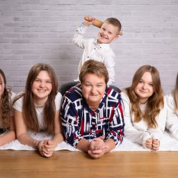 Pan Aparacik Dawid Kubica - Portret rodzinny: kobieta w koszuli w kratę leży na dywanie z czterema nastolatkami, chłopiec stoi za nią z wałkiem w ręku, uśmiechnięci, tło z cegły.