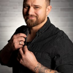 Pan Aparacik Dawid Kubica - Portret uśmiechniętego mężczyzny z brodą, w czarnej koszuli w kropki, z tatuażem na przedramieniu i obrączką na palcu, pozującego na tle ściany z cegieł.