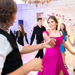 Pan Aparacik Dawid Kubica - Grupa młodych ludzi w eleganckich strojach tańczy w jasnym, udekorowanym wnętrzu podczas uroczystości. Dziewczyna w fuksjowej sukience uśmiecha się do partnera.