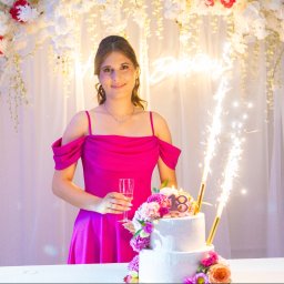 Pan Aparacik Dawid Kubica - Uśmiechnięta brunetka w różowej sukience, trzyma kieliszek szampana obok tortu na 18 urodziny, dekoracje kwiatowe i płonące fontanny.