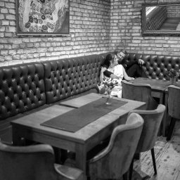 Pan Aparacik Dawid Kubica - Czarno-białe zdjęcie pary w restauracji z ceglanymi ścianami, siedzącej na skórzanej kanapie. Na stole wazon z kwiatami. Klimatyczne oświetlenie, obraz na ścianie.