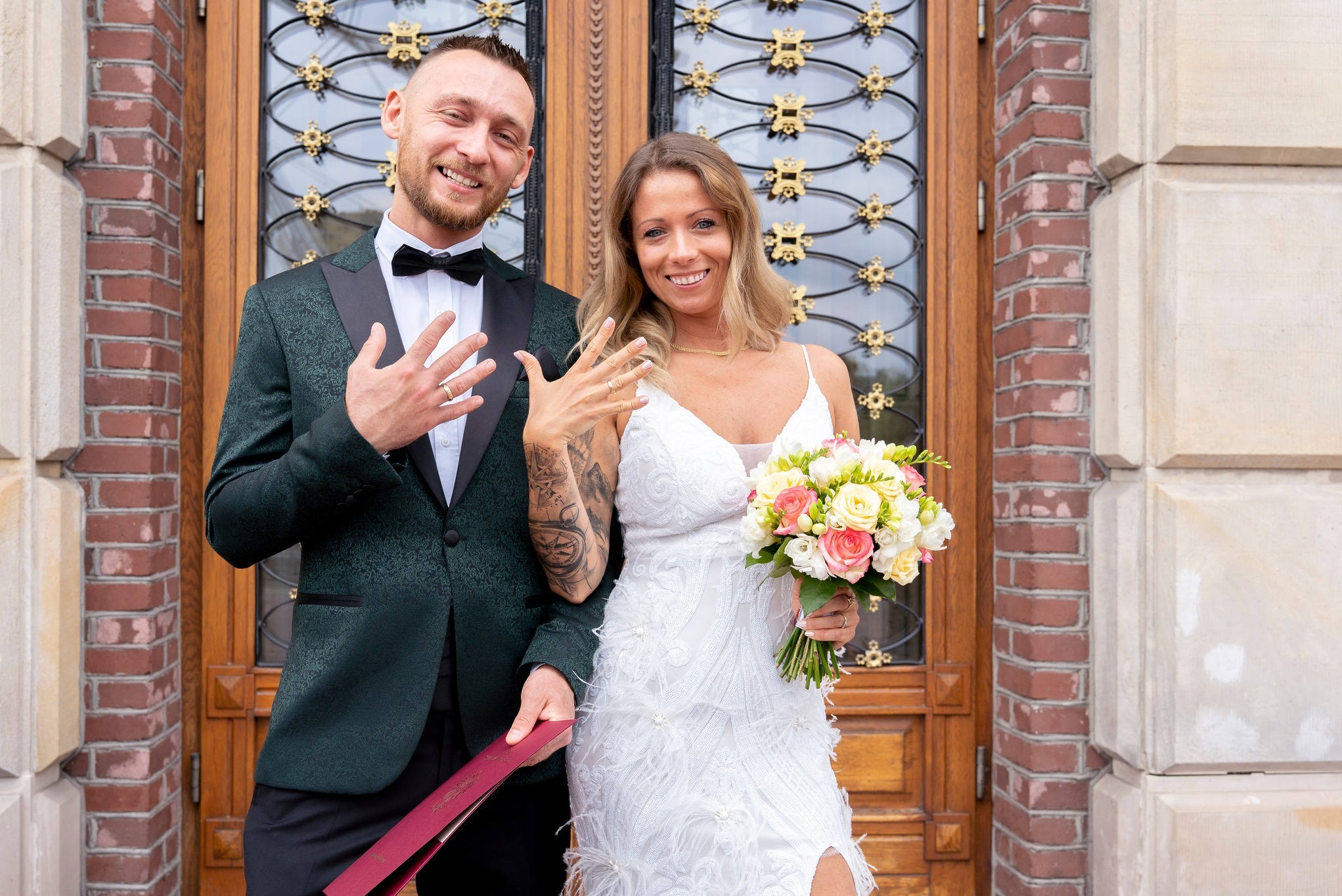 Uśmiechnięta para młoda prezentuje obrączki na tle zdobionych drzwi i ceglanej ściany. Panna młoda w białej sukni z bukietem, pan młody w zielonym smokingu z muchą.