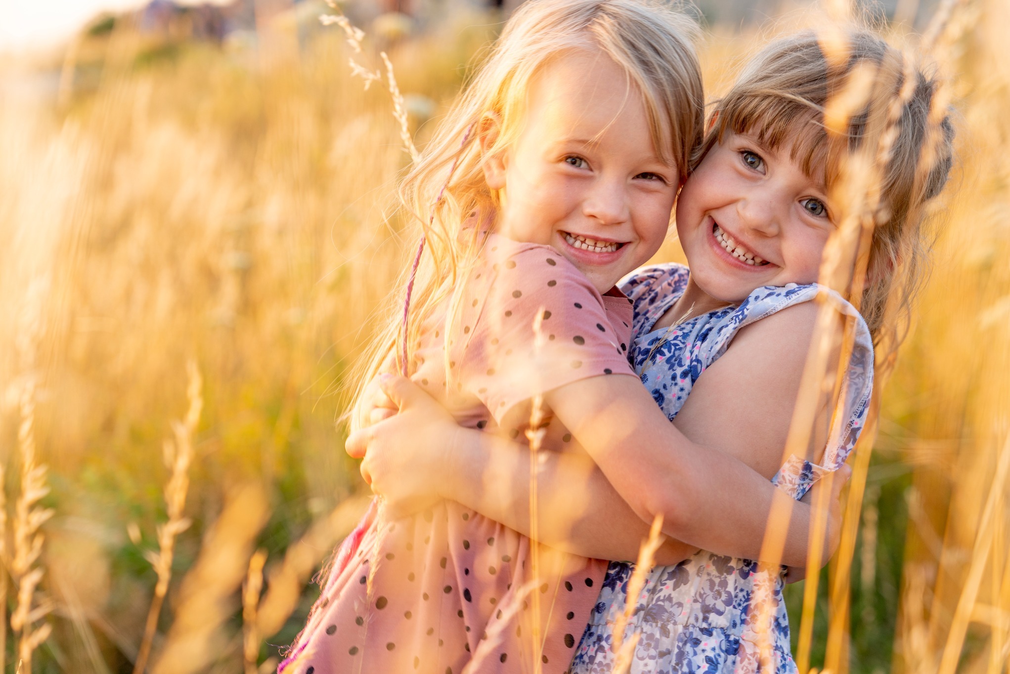 Dwie uśmiechnięte dziewczynki obejmują się na tle pola zboża w ciepłym, zachodzącym słońcu, jedna w różowej sukience w kropki, druga w niebieskiej w kwiaty.