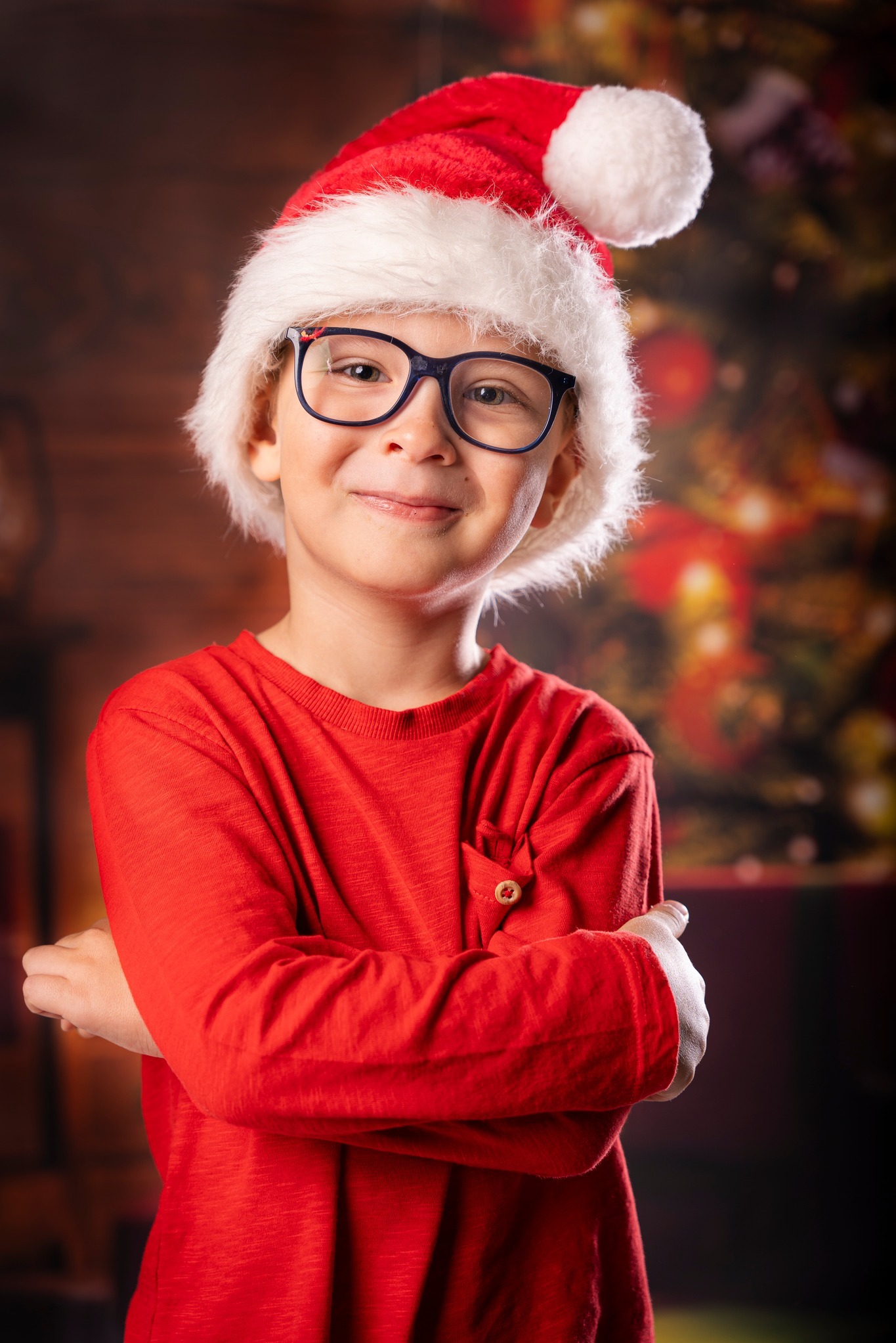 Uśmiechnięty chłopiec w okularach i czerwonym swetrze, ubrany w czapkę Świętego Mikołaja z puszystym pomponem, pozuje na tle rozświetlonej choinki.