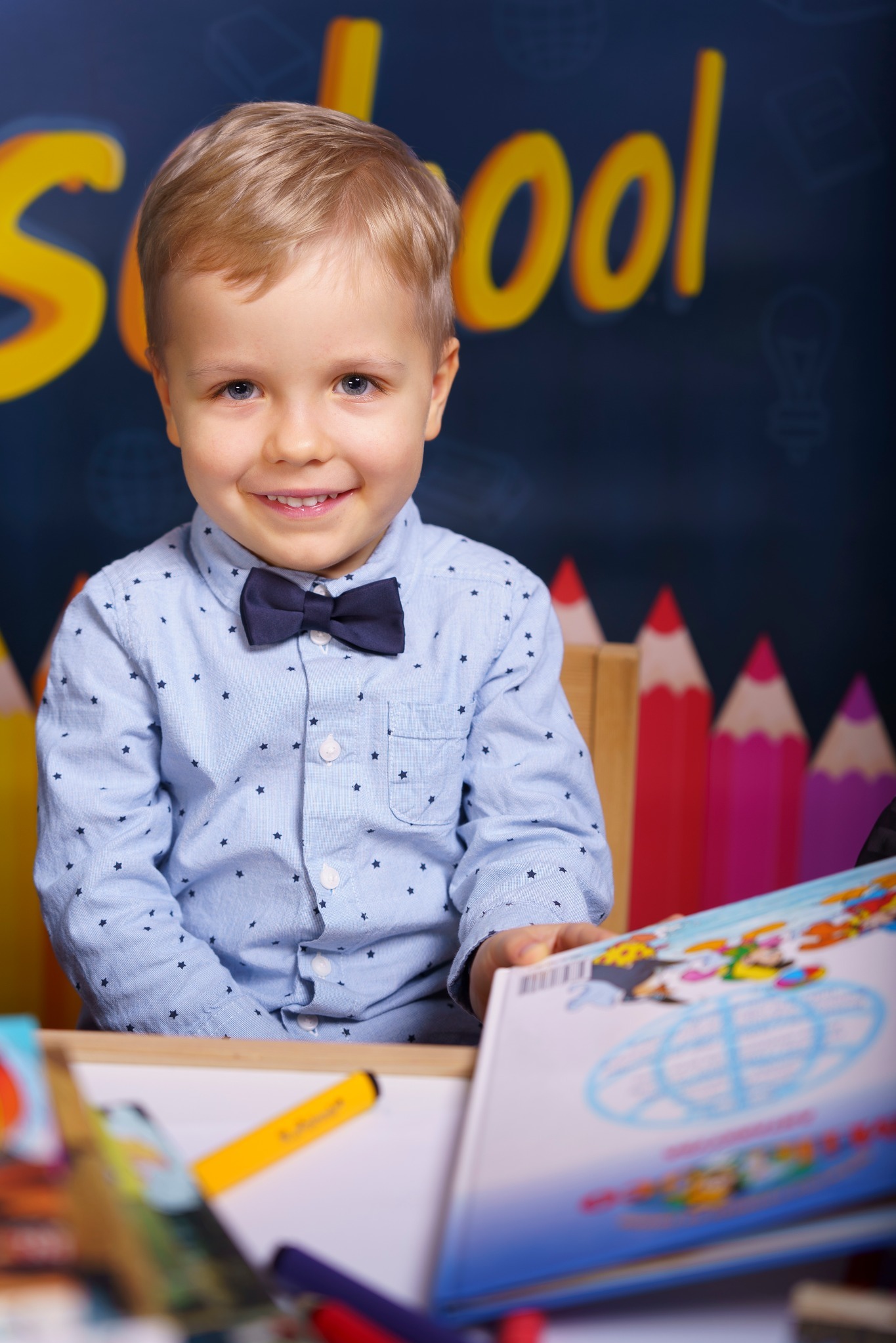Uśmiechnięty blond chłopiec w koszuli w gwiazdki i granatowej muszce siedzi za biurkiem z książkami i flamastrami, na tle tablicy z napisem 'School' i kolorowych kredek.