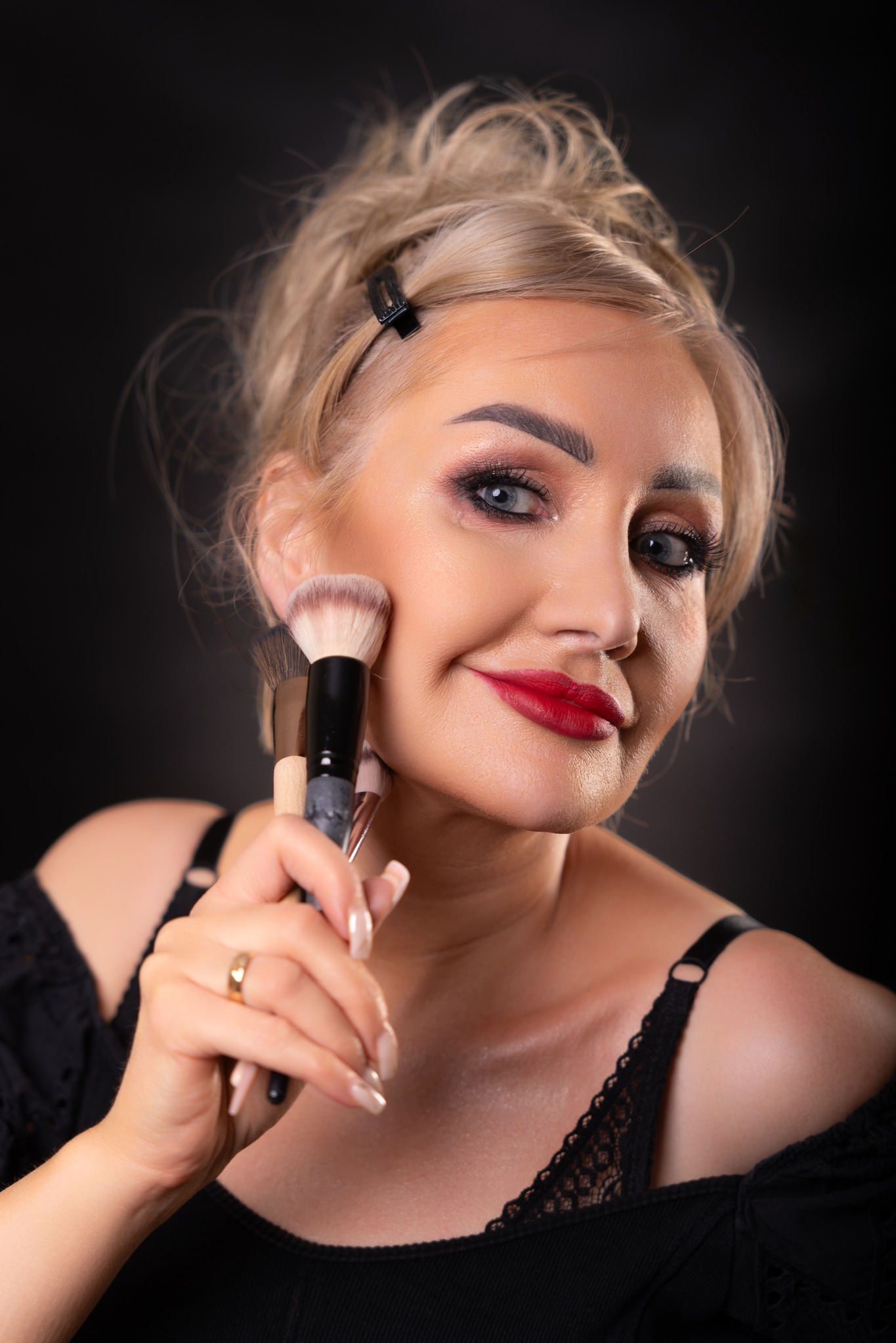 Uśmiechnięta blondynka z wyrazistym makijażem oczu i czerwonymi ustami trzymająca pędzle do makijażu przy policzku, na czarnym tle.