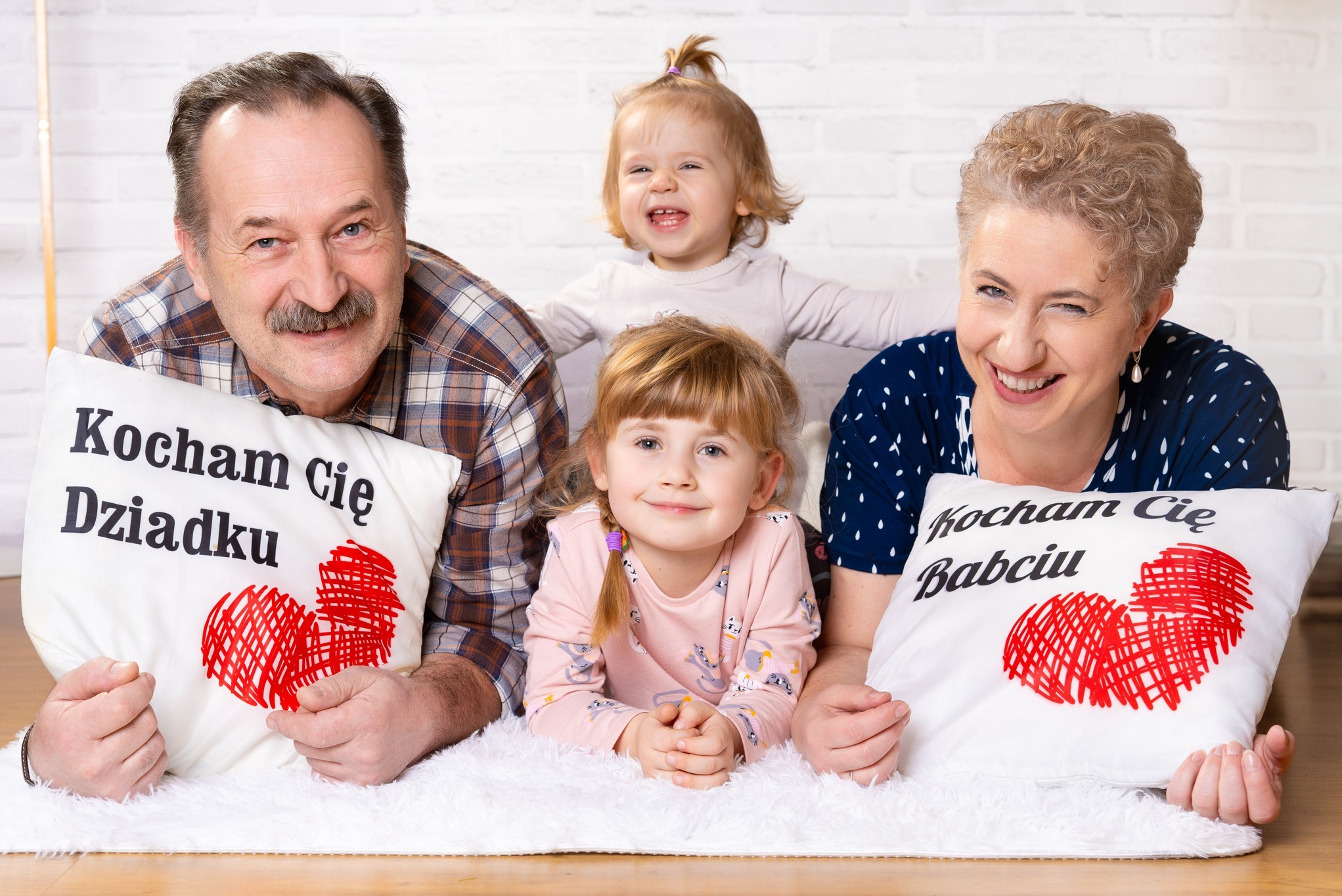 Uśmiechnięta rodzina: dziadek w koszuli w kratę z poduszką 'Kocham Cię Dziadku', babcia mrugająca okiem z poduszką 'Kocham Cię Babciu' oraz dwie wnuczki na białym puszystym dywanie na tle białej...