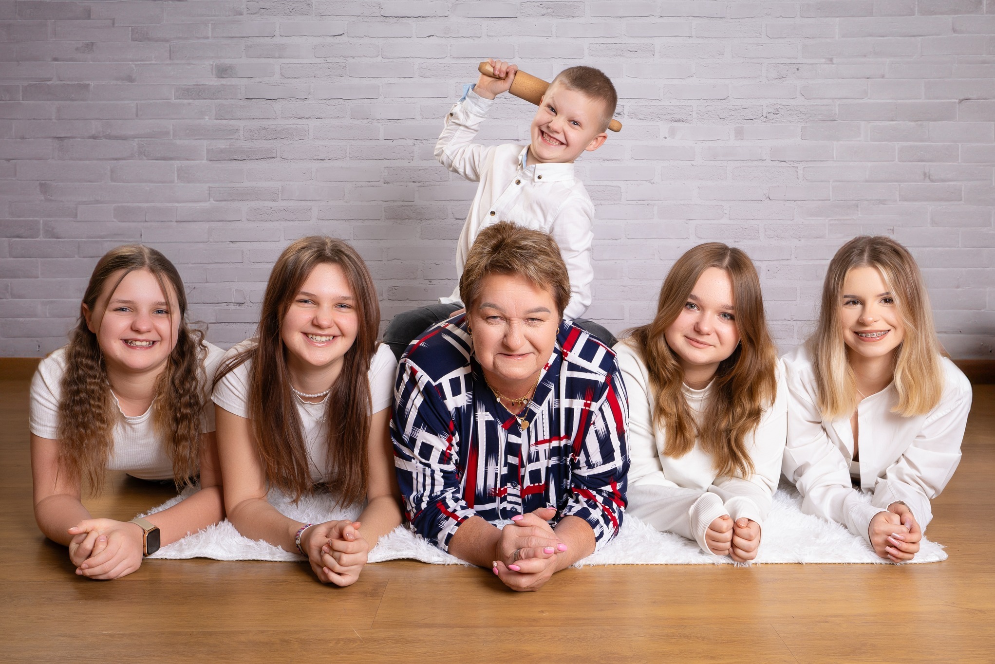 Portret rodzinny: kobieta w koszuli w kratę leży na dywanie z czterema nastolatkami, chłopiec stoi za nią z wałkiem w ręku, uśmiechnięci, tło z cegły.
