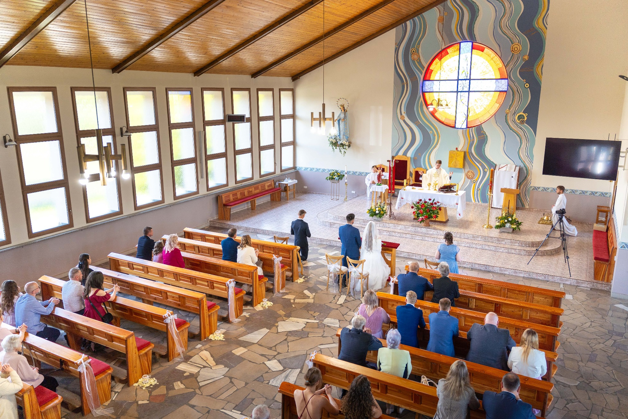Widok z góry na kościół podczas ceremonii ślubnej. Para młoda stoi przed ołtarzem, a goście siedzą w ławkach udekorowanych białymi wstążkami i płatkami kwiatów.