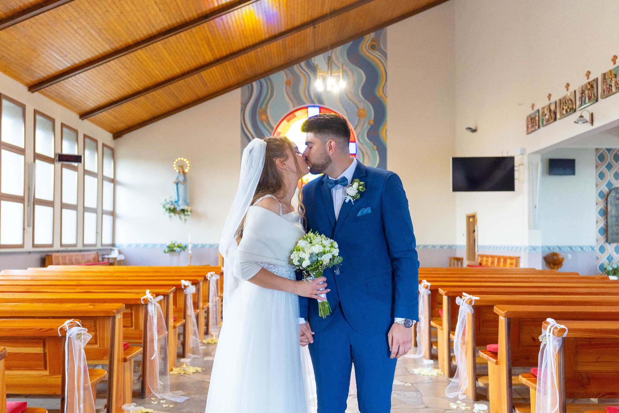 Pocałunek młodej pary w kościele, on w błękitnym garniturze z muszką, ona w białej sukni z welonem i bukietem. Dekorowane ławki, witrażowe okno w tle.