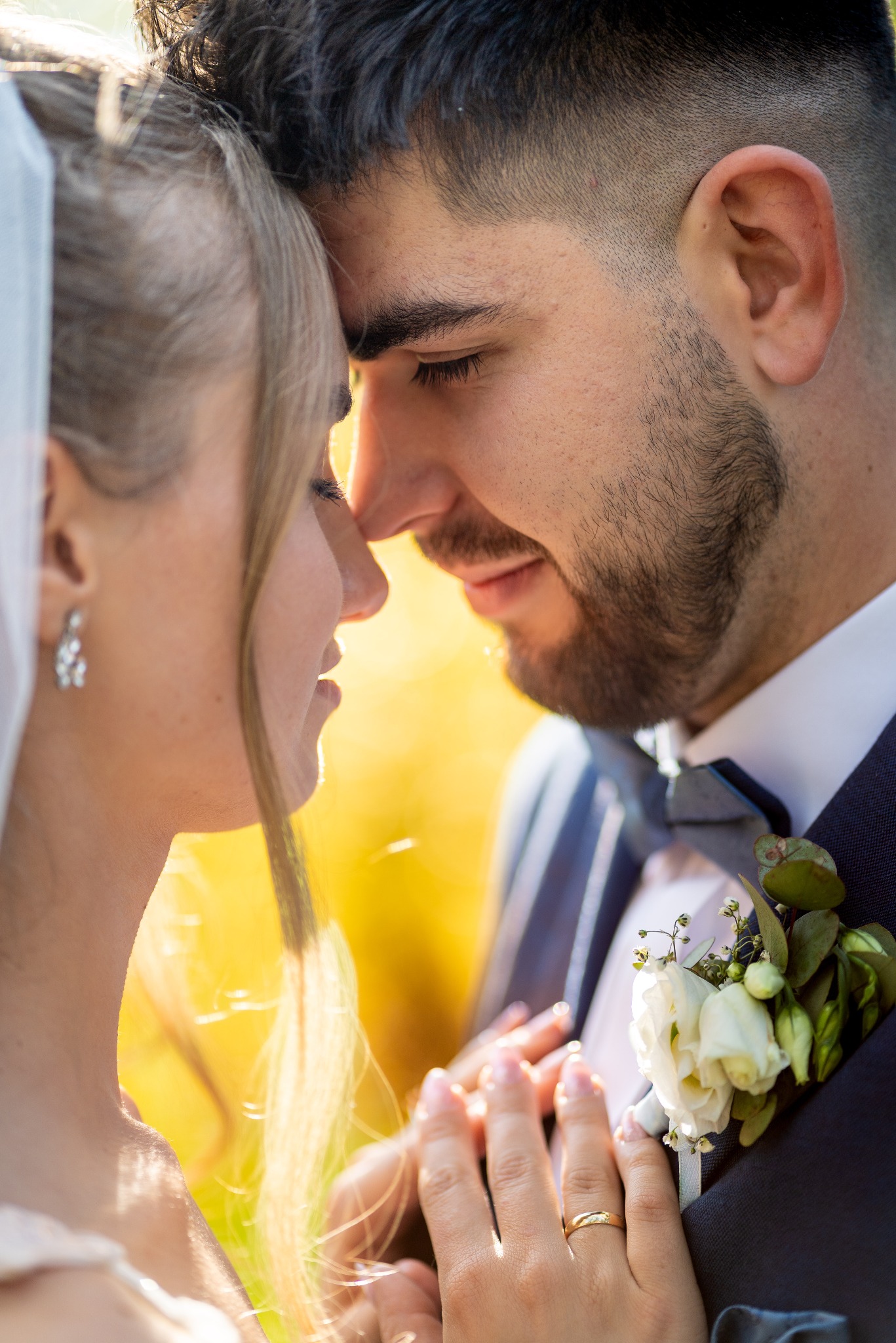 Zbliżenie na parę młodą w czułym uścisku, z delikatnym bukiecikiem w butonierce pana młodego i welonem panny młodej. Ciepłe, naturalne światło podkreśla intymność chwili.