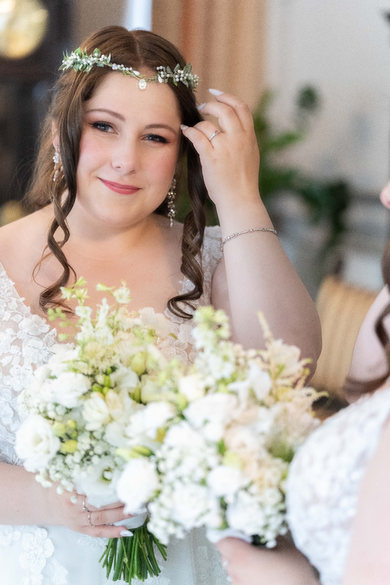 Uśmiechnięta panna młoda z kwiatami, w koronkowej sukni i wianku, poprawia włosy, widoczna w odbiciu lustra. Delikatny makijaż i biżuteria.