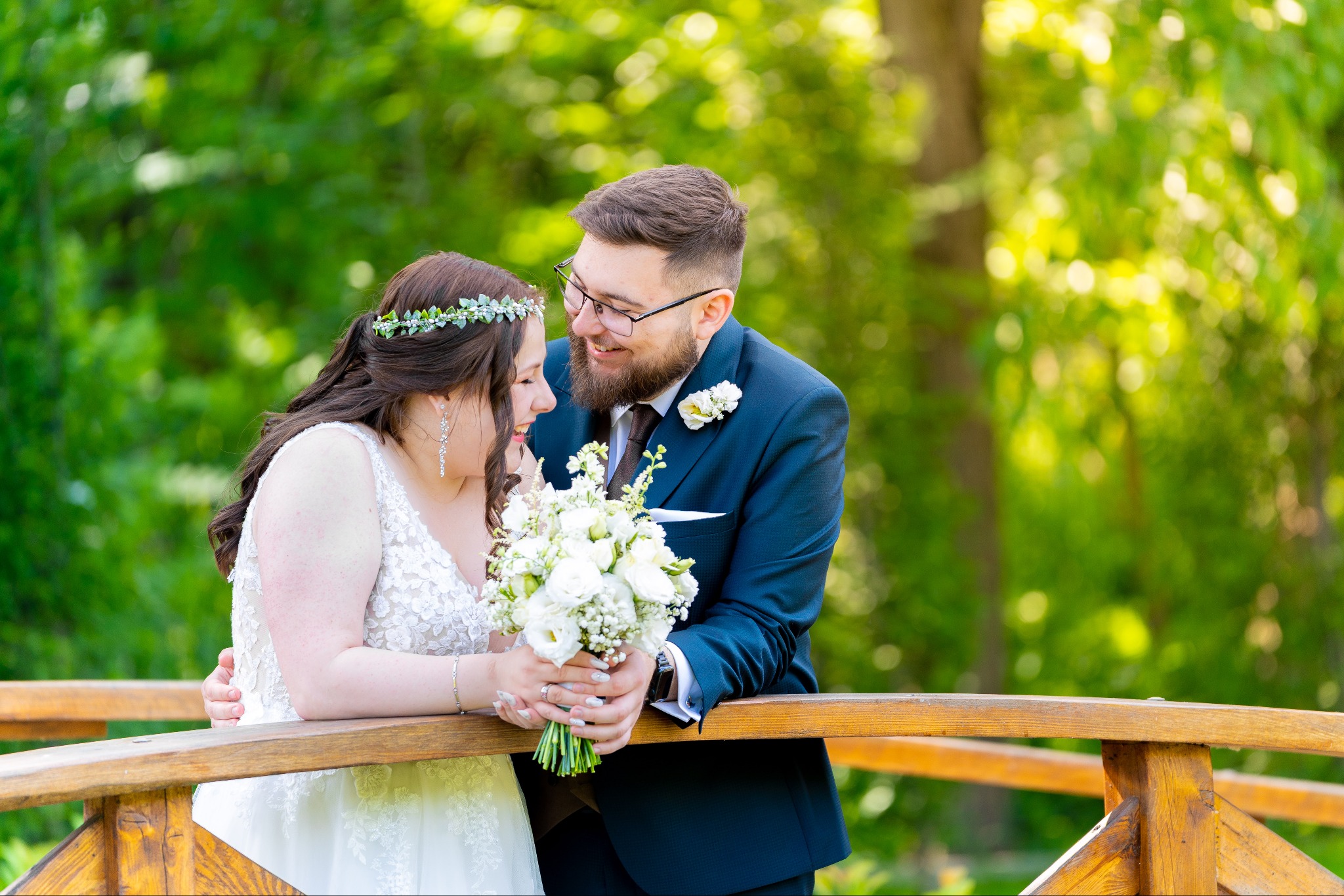 Uśmiechnięta para młoda na drewnianym moście w zielonym plenerze. Panna młoda w sukni z koronką i wiankiem, pan młody w granatowym garniturze.