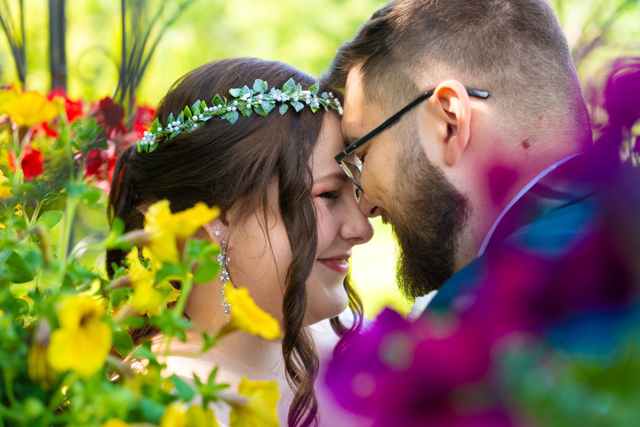 Uśmiechnięta para młoda, panna młoda w wianku z kwiatów i długich kolczykach, delikatnie dotykają się czołami, otoczeni kolorowymi kwiatami w słoneczny dzień.