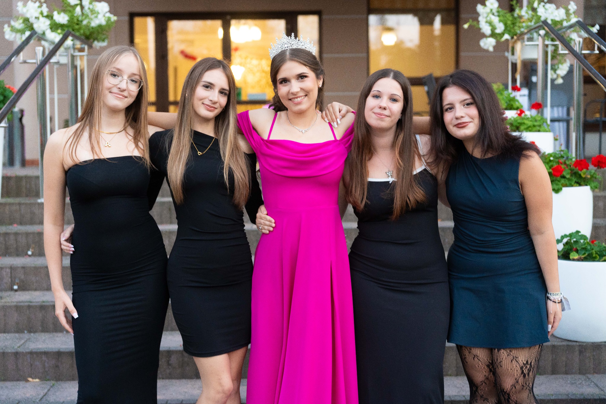 Grupa pięciu młodych kobiet w sukienkach pozuje na schodach. Jedna z nich ma na głowie diadem. W tle widoczne kwiaty w donicach i budynek.