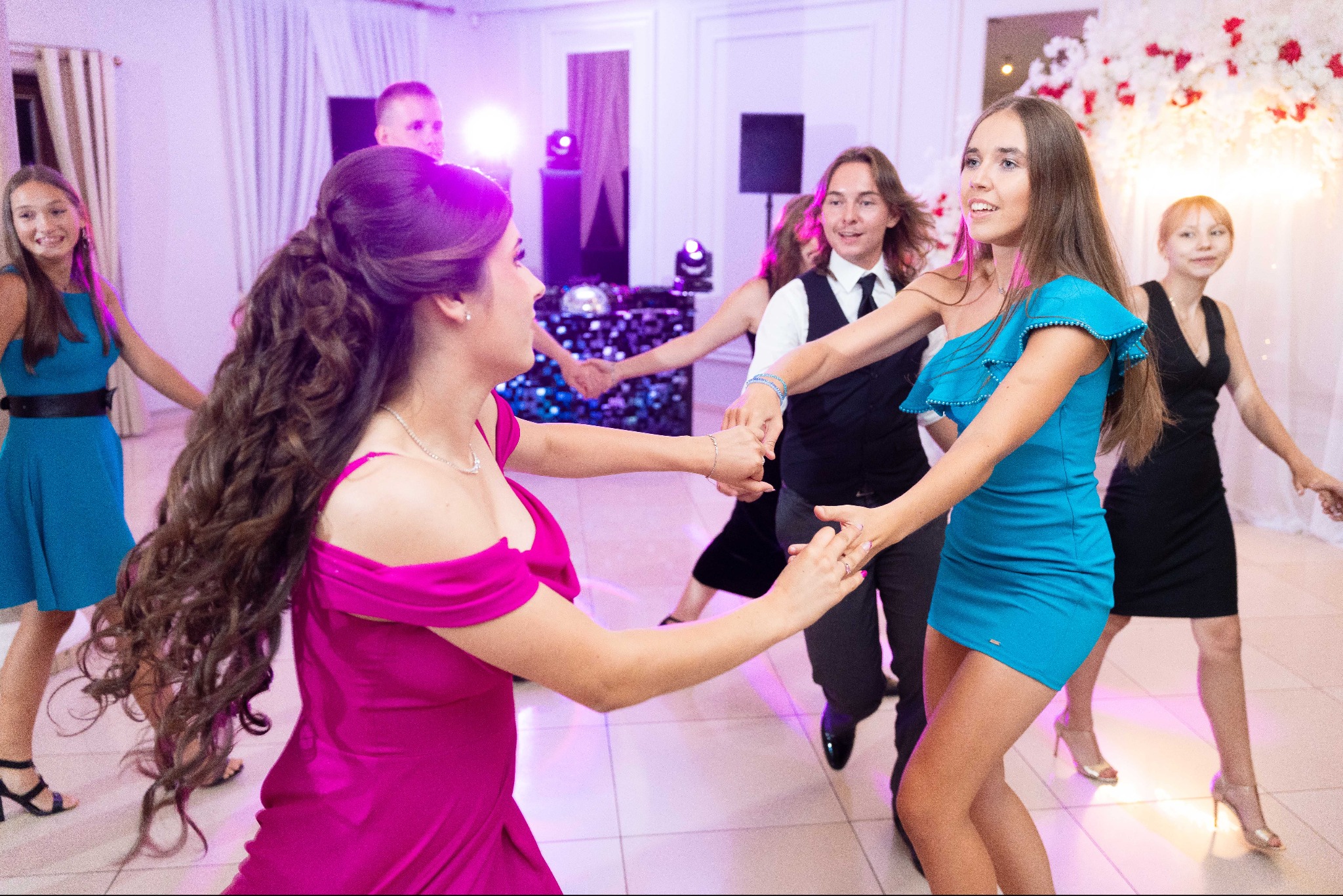 Roztańczona grupa młodych ludzi w eleganckich strojach, bawiąca się na parkiecie podczas imprezy. W tle dekoracje kwiatowe i efekty świetlne.