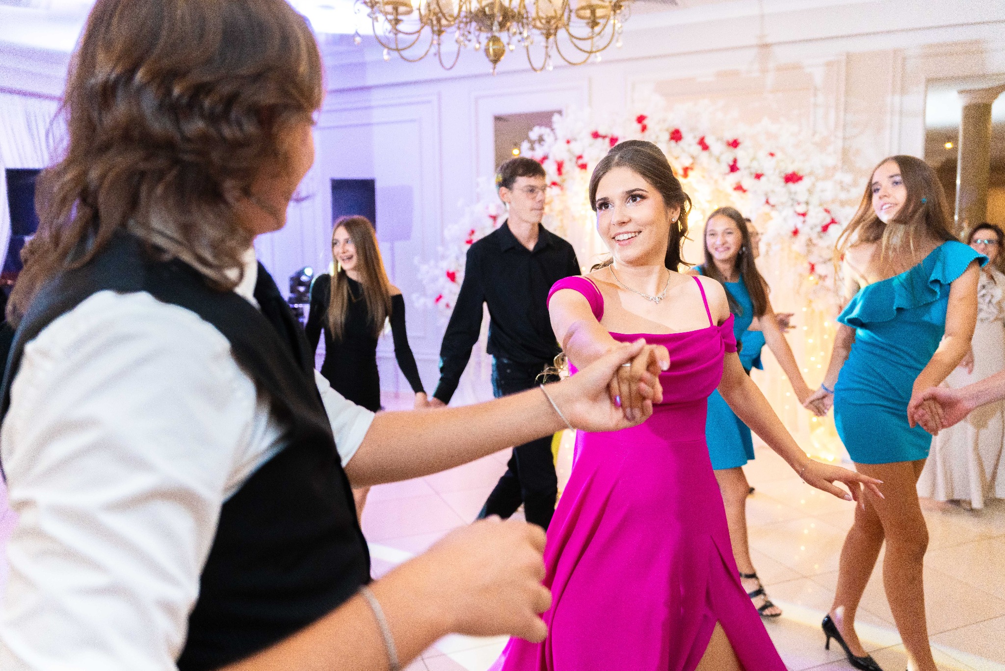 Grupa młodych ludzi w eleganckich strojach tańczy w jasnym, udekorowanym wnętrzu podczas uroczystości. Dziewczyna w fuksjowej sukience uśmiecha się do partnera.