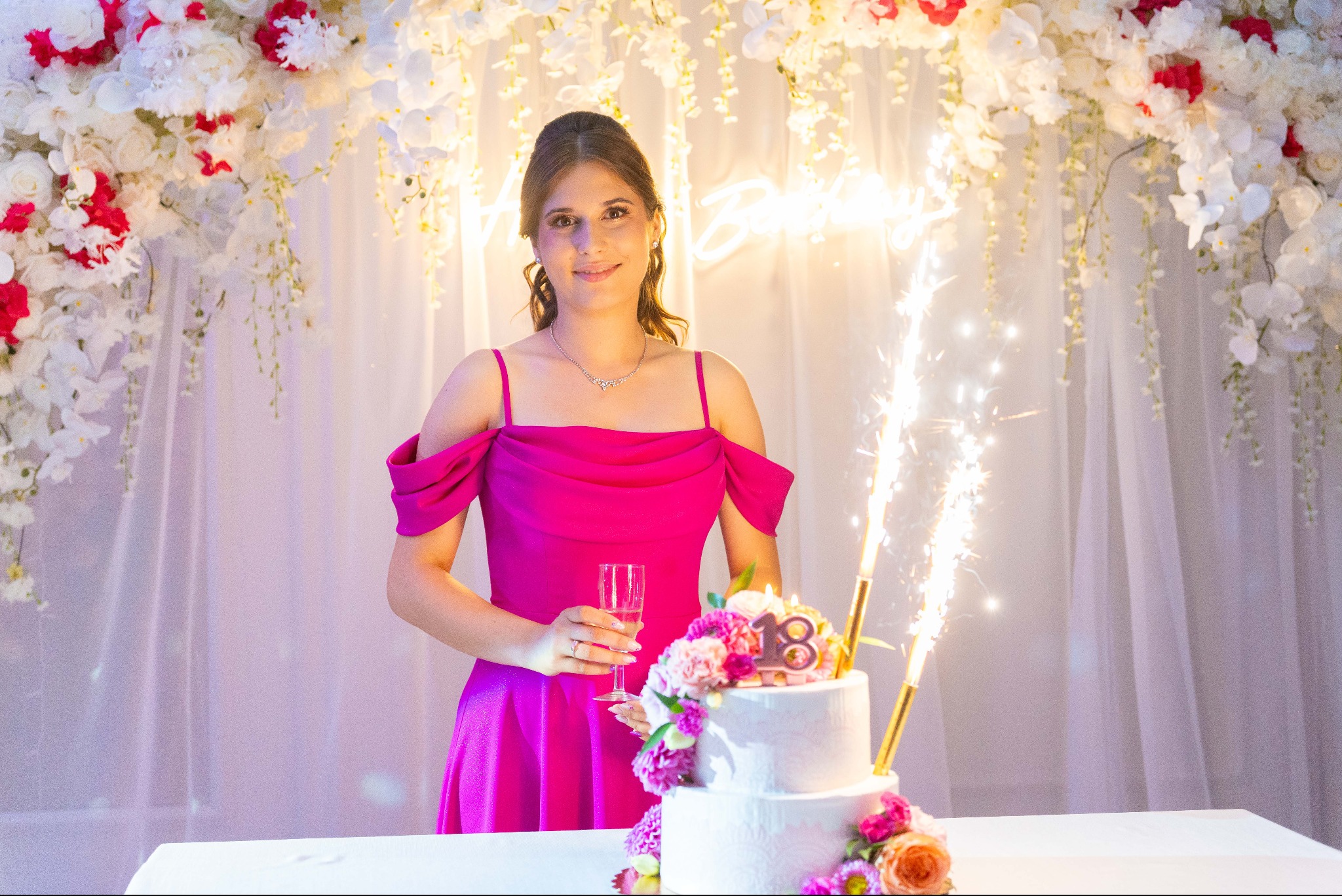 Uśmiechnięta brunetka w różowej sukience, trzyma kieliszek szampana obok tortu na 18 urodziny, dekoracje kwiatowe i płonące fontanny.