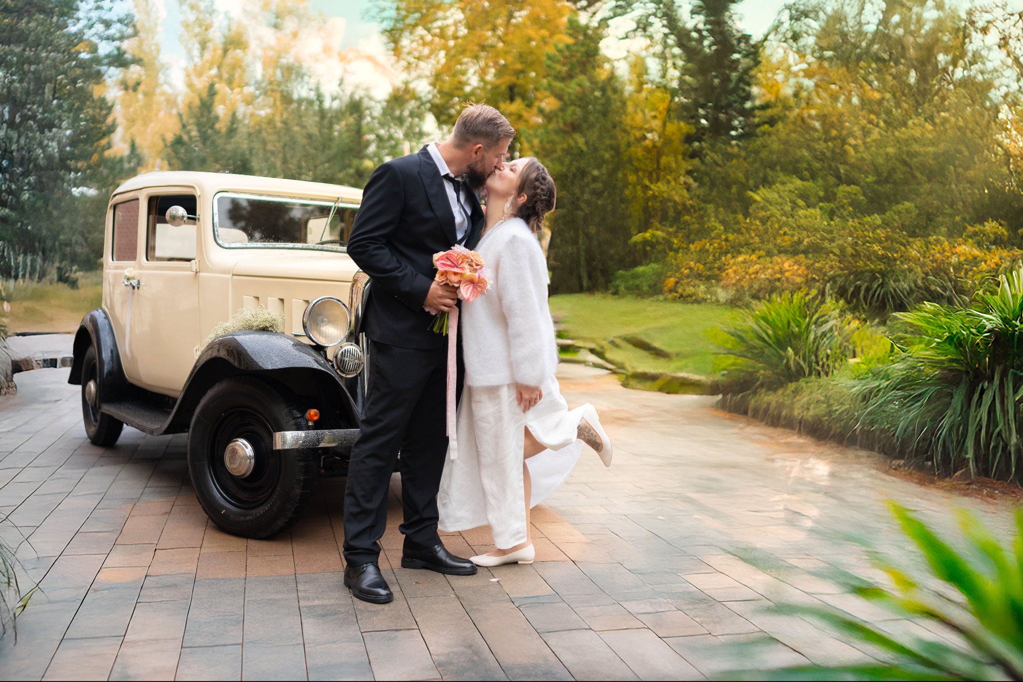 Pocałunek pary młodej w Siewierzu na tle beżowego auta retro i zieleni. Panna młoda w białym stroju, pan młody w czarnym garniturze. Romantyczna sesja plenerowa.
