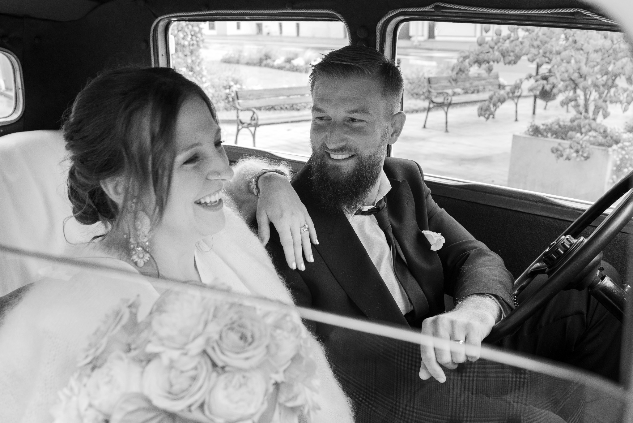 Czarno-białe zdjęcie uśmiechniętej pary młodej siedzącej w zabytkowym samochodzie. Panna młoda z bukietem i kolczykami, pan młody w garniturze z butonierką.