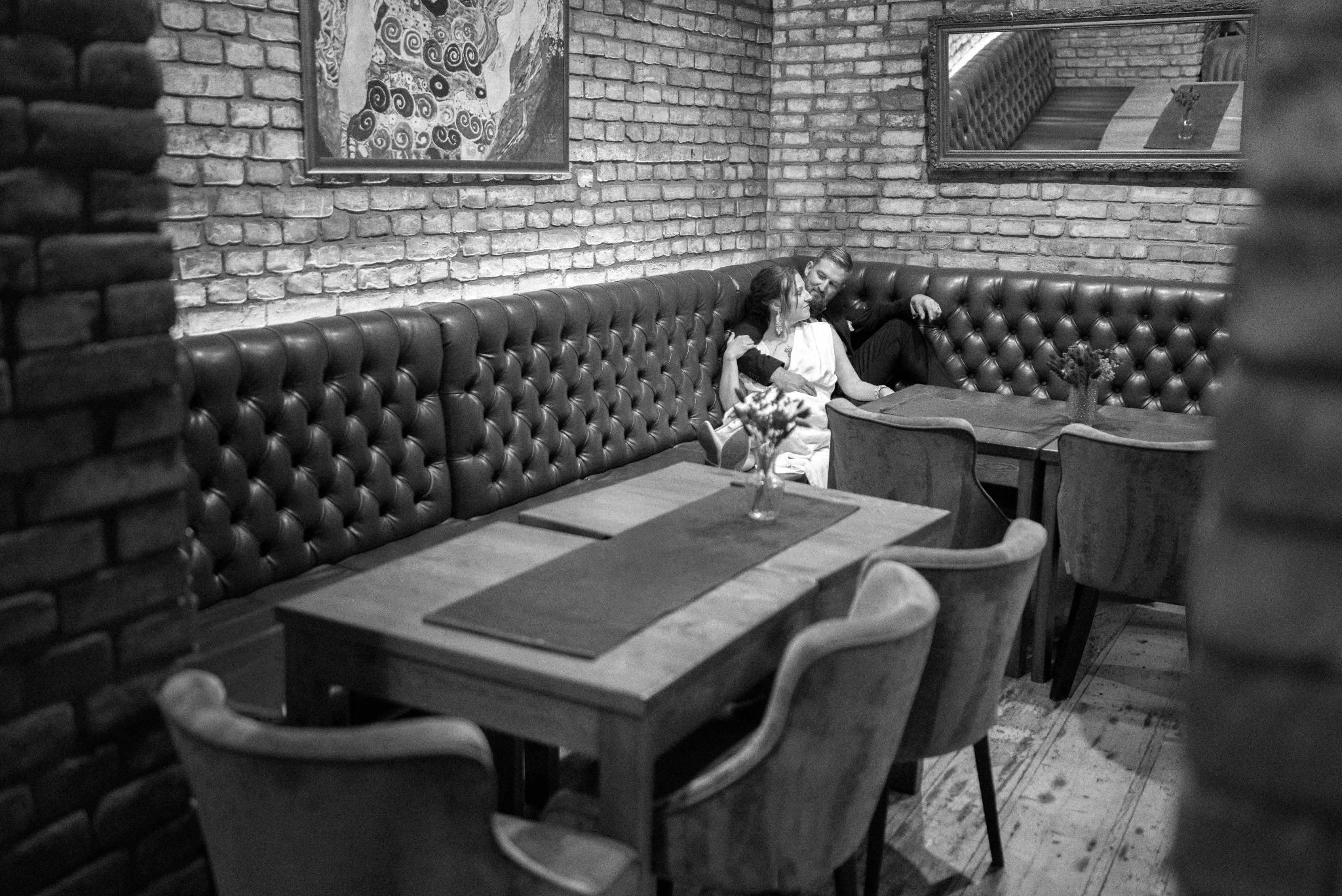 Czarno-białe zdjęcie pary w restauracji z ceglanymi ścianami, siedzącej na skórzanej kanapie. Na stole wazon z kwiatami. Klimatyczne oświetlenie, obraz na ścianie.