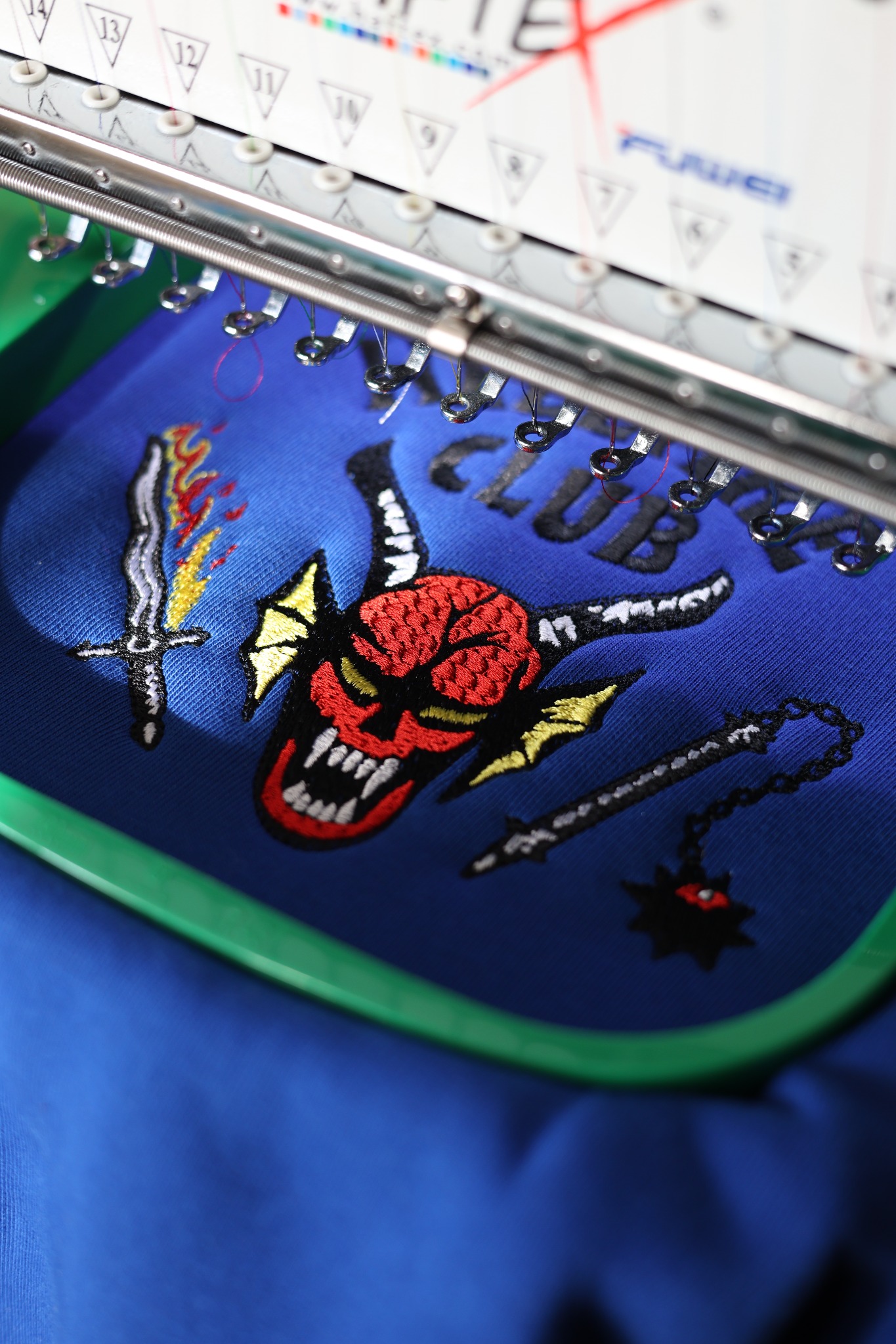 Proces wykonywania haftu na niebieskiej tkaninie, widoczny motyw czaszki z rogami, miecza i maczugi z kolcami, igły maszyny do haftowania w trakcie pracy.