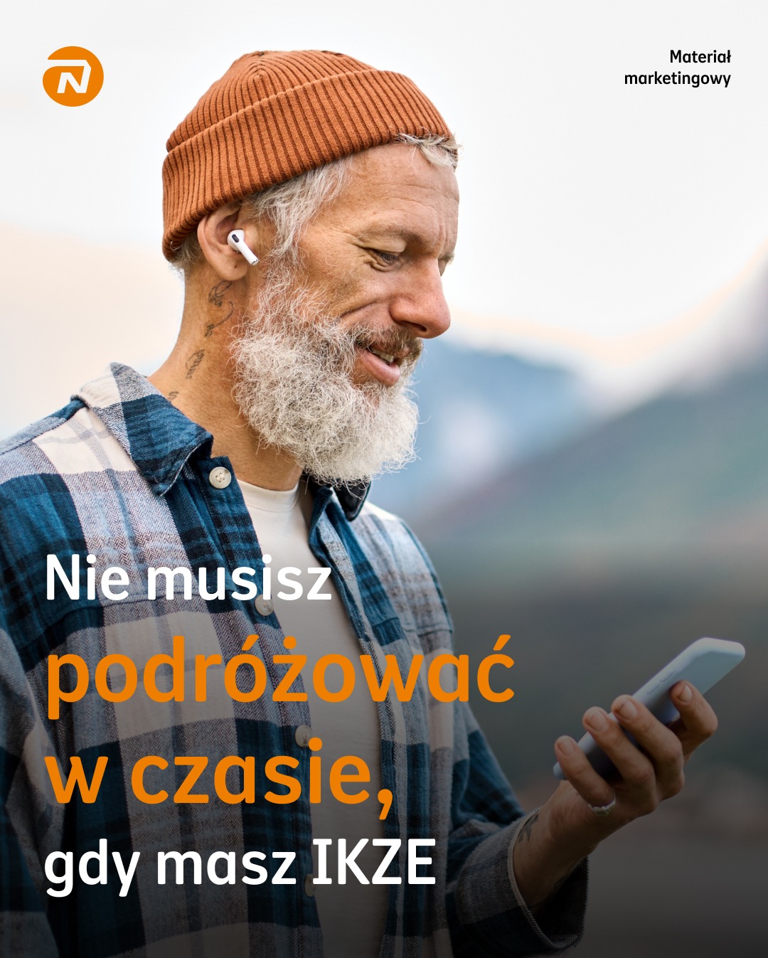 Uśmiechnięty mężczyzna z brodą w czapce, z telefonem w dłoni, na tle gór. Reklama IKZE z tekstem 'Nie musisz podróżować w czasie, gdy masz IKZE'.