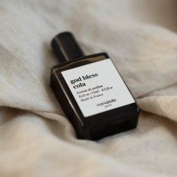 suzanephoto - Butelka perfum 'God Bless Cola' Versatile Paris na beżowej, pogniecionej tkaninie, z widocznym napisem 'Extrait de parfum Roll-on / 15ml - 0.5 fl.oz Made in France'.