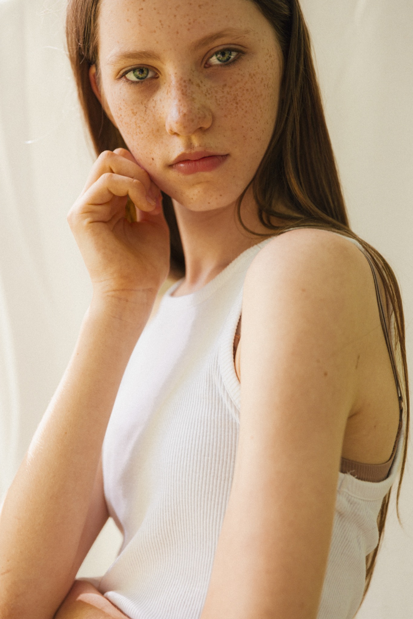 Młoda kobieta z piegami, zielonymi oczami i długimi brązowymi włosami, ubrana w białą koszulkę na ramiączkach, pozująca z ręką przy twarzy na jasnym tle.