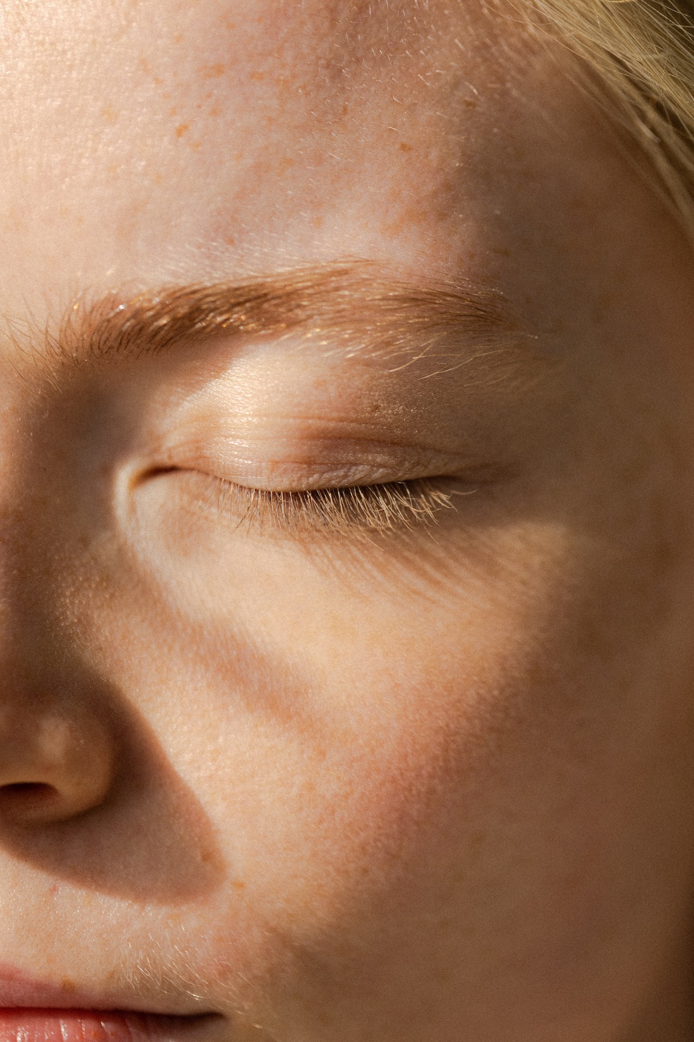 Portret zbliżeniowy zamkniętego oka kobiety z jasną cerą i piegami, oświetlonego naturalnym światłem, ukazujący teksturę skóry i delikatne rzęsy.