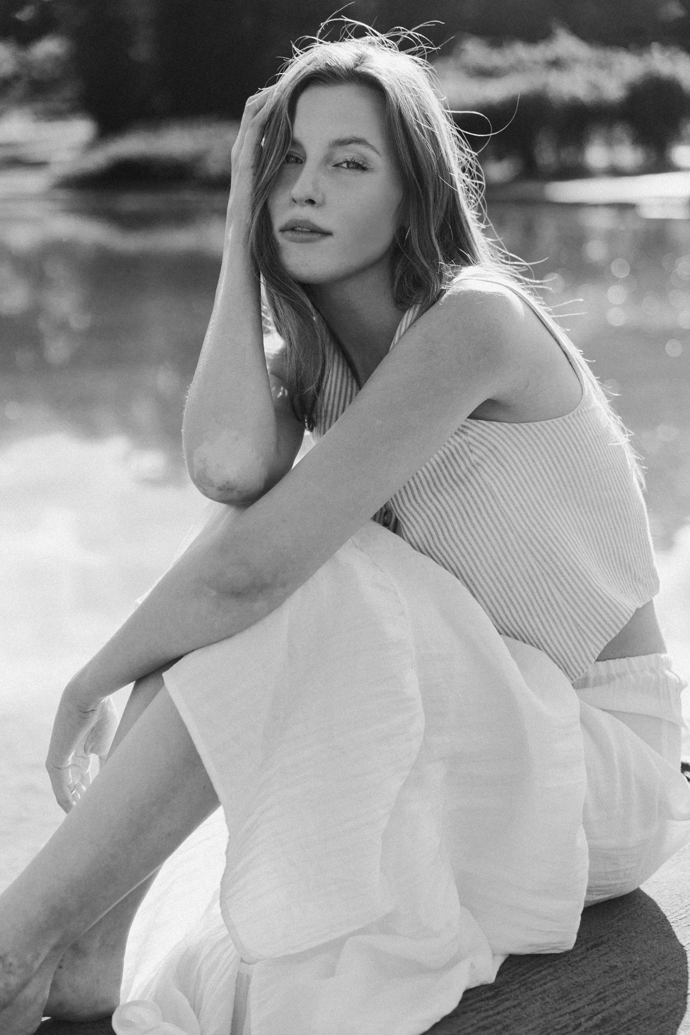 Portret w czerni i bieli: młoda kobieta siedzi z ręką na głowie, w tle rozmyte jezioro i drzewa.