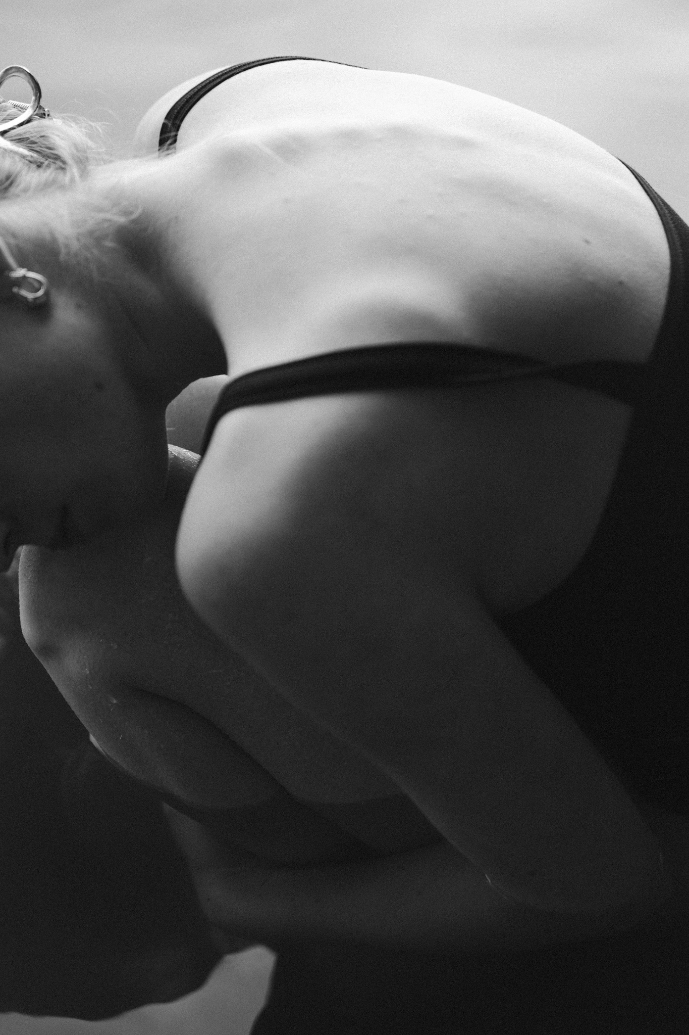 Czarno-białe, artystyczne ujęcie pochylonej kobiety z widocznymi plecami, ramieniem i fragmentem włosów spiętych ozdobną spinką.