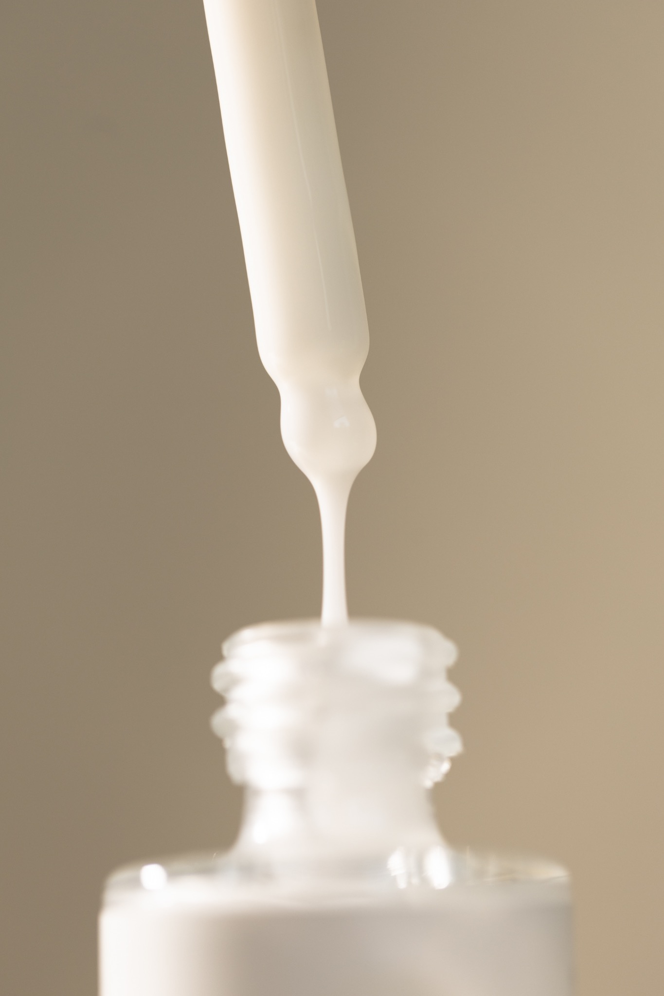 Kropla białego serum z pipety opada do butelki, delikatne tło w odcieniach beżu.