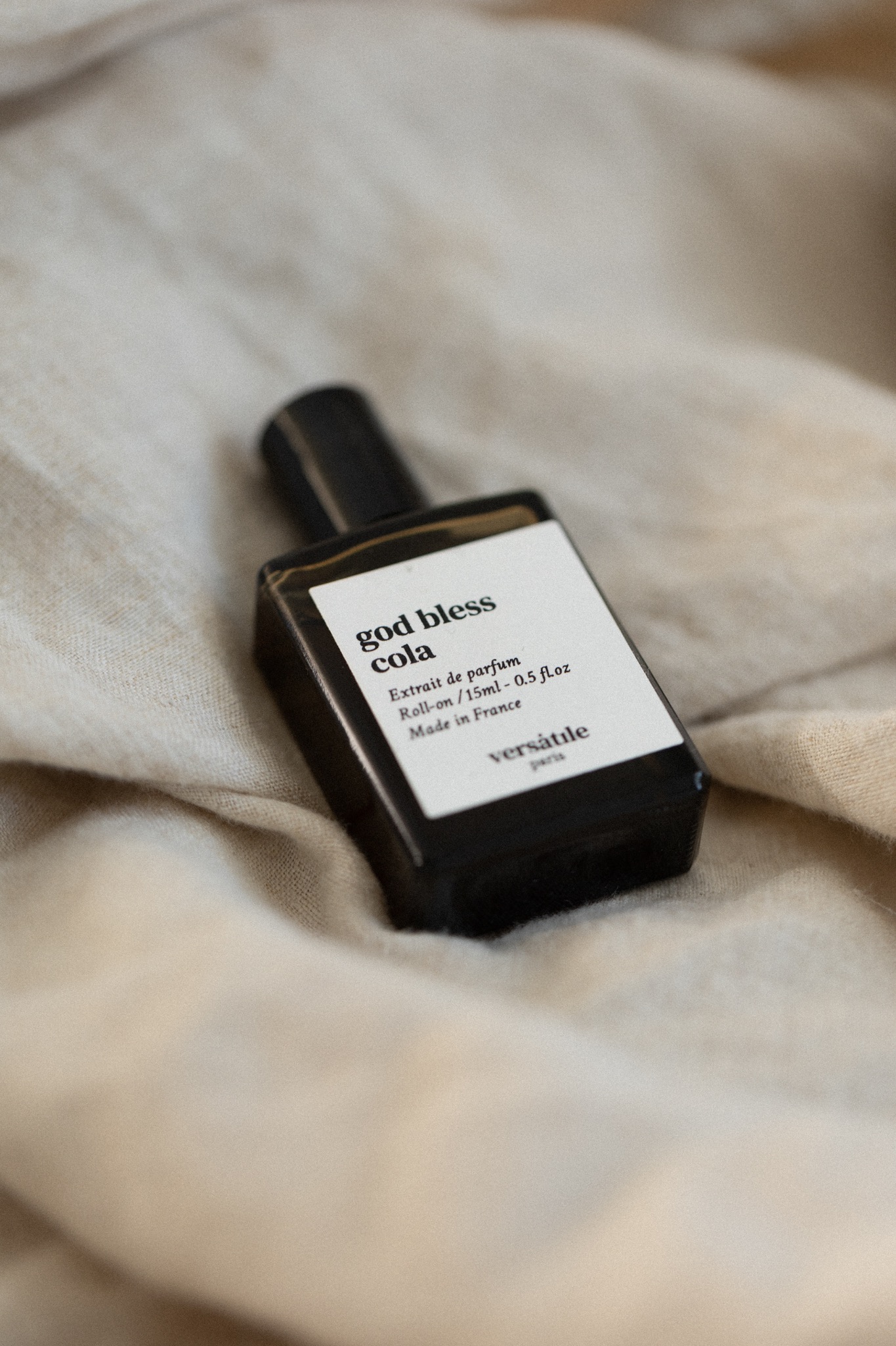 Butelka perfum 'God Bless Cola' Versatile Paris na beżowej, pogniecionej tkaninie, z widocznym napisem 'Extrait de parfum Roll-on / 15ml - 0.5 fl.oz Made in France'.
