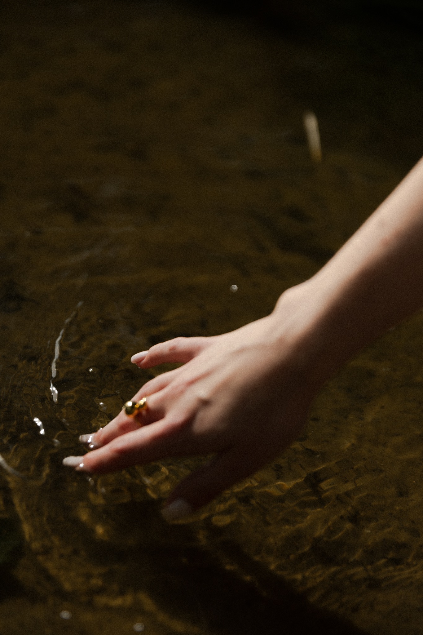 Dłoń kobiety z dużym złotym pierścieniem dotyka powierzchni wody, tworząc delikatne fale i refleksy świetlne.