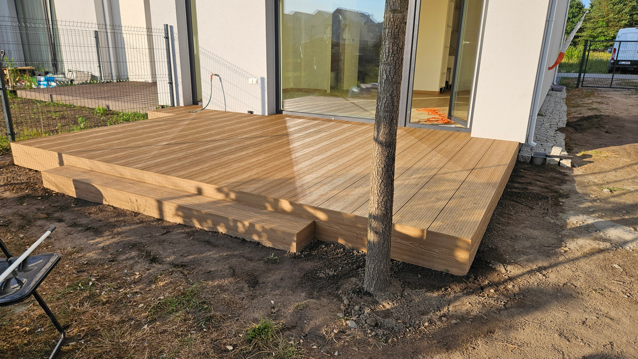 Nowoczesny taras kompozytowy z dwustopniowymi schodami, ułożony pod kątem, z pniem drzewa wystającym przez deski, na tle białego budynku i zielonego ogrodu.