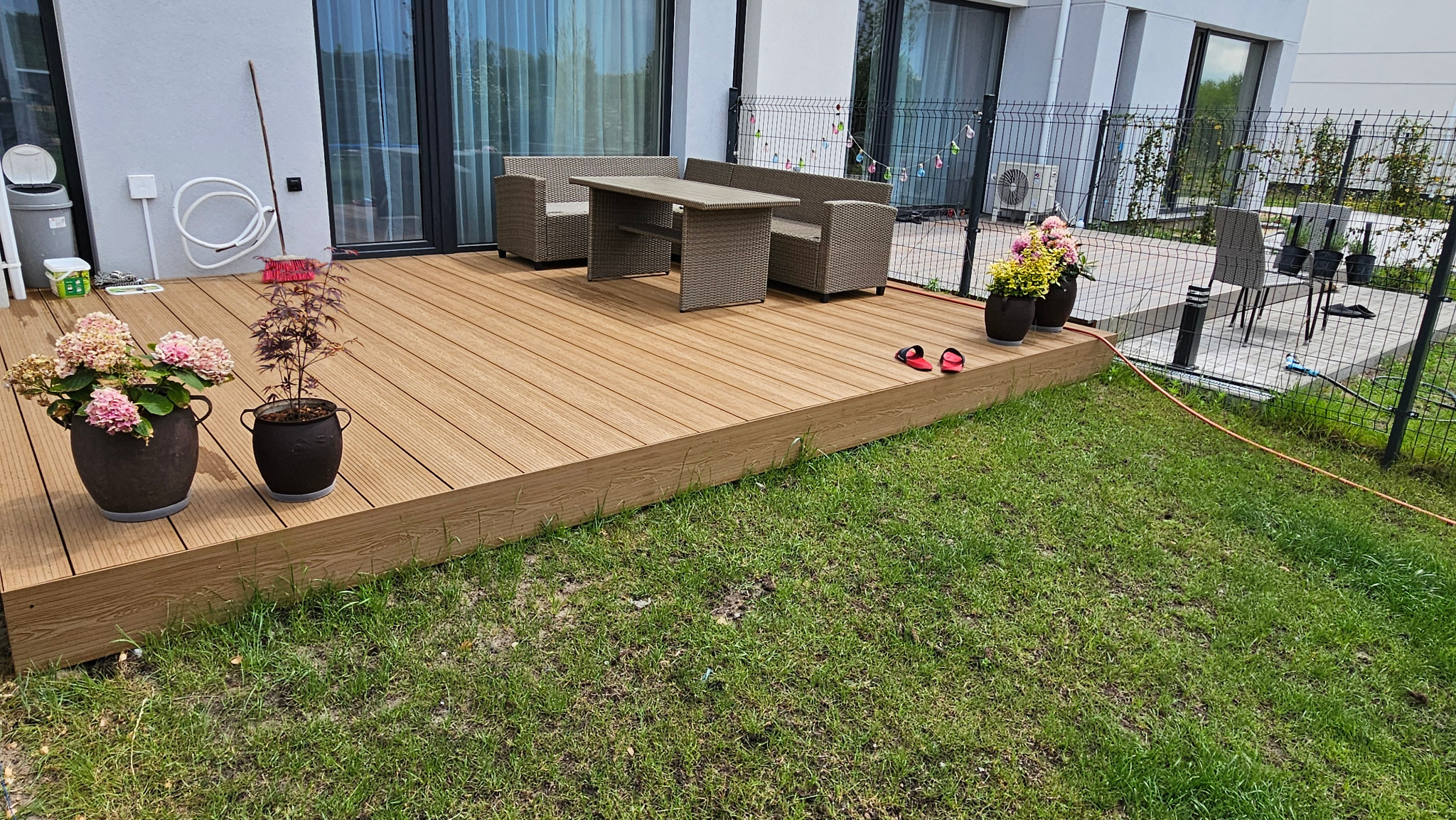 Jasny taras z desek kompozytowych z meblami ogrodowymi z technorattanu, doniczkami z kwiatami oraz parą czerwonych klapek na obrzeżu, widok z poziomu trawnika.