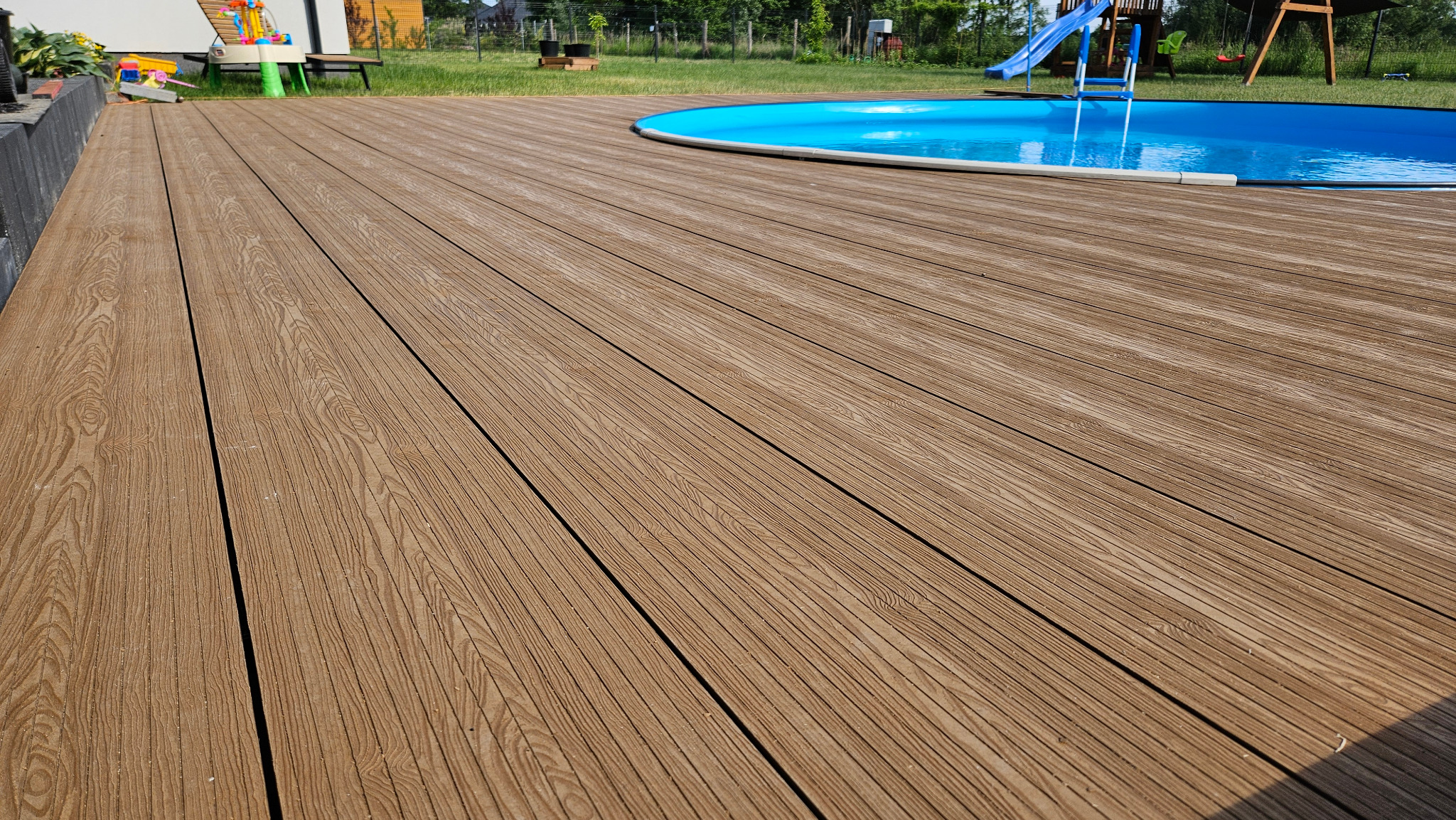 Nowoczesny taras z desek kompozytowych w odcieniach brązu, otaczający okrągły basen na zielonym trawniku.