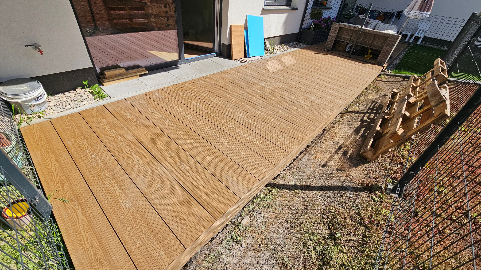Jasnobrązowy taras z desek kompozytowych, widok z góry, częściowo ukończony, z paletą i elementami montażowymi obok, przy budynku.