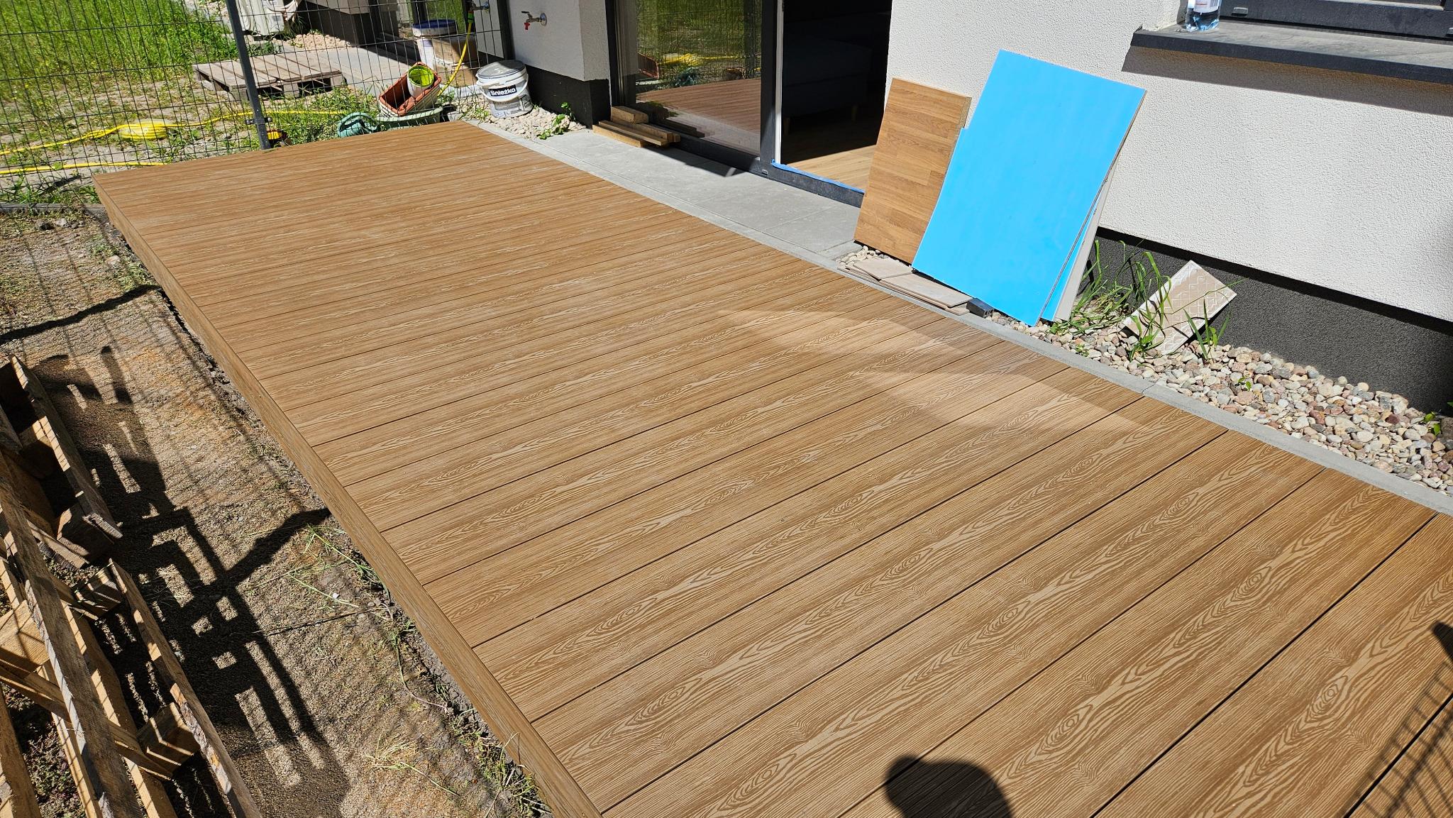 Nowo wybudowany taras z desek kompozytowych, widok z góry, z elementami wykończeniowymi opartymi o ścianę budynku.