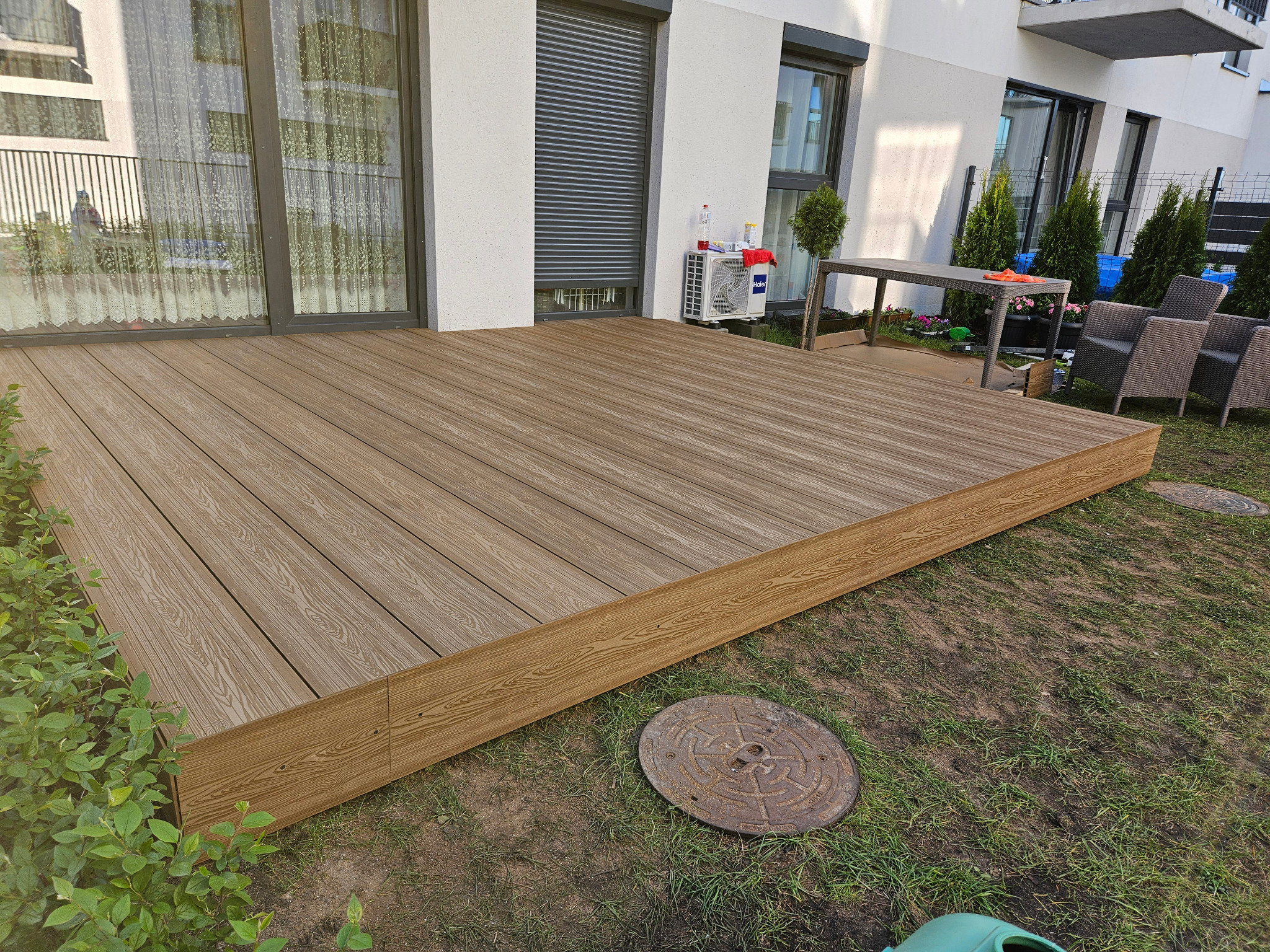 Nowoczesny taras z desek kompozytowych w odcieniach brązu, przylegający do budynku z dużymi oknami i roletami zewnętrznymi, widoczne elementy wyposażenia ogrodu i zieleń.