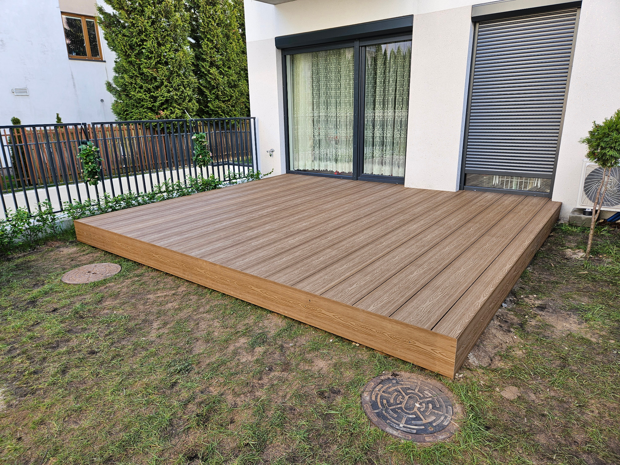 Nowoczesny taras z desek kompozytowych o ciepłym, brązowym odcieniu, przylegający do białego domu z oknami przesuwnymi i roletami zewnętrznymi, otoczony trawnikiem z widocznymi studzienkami...