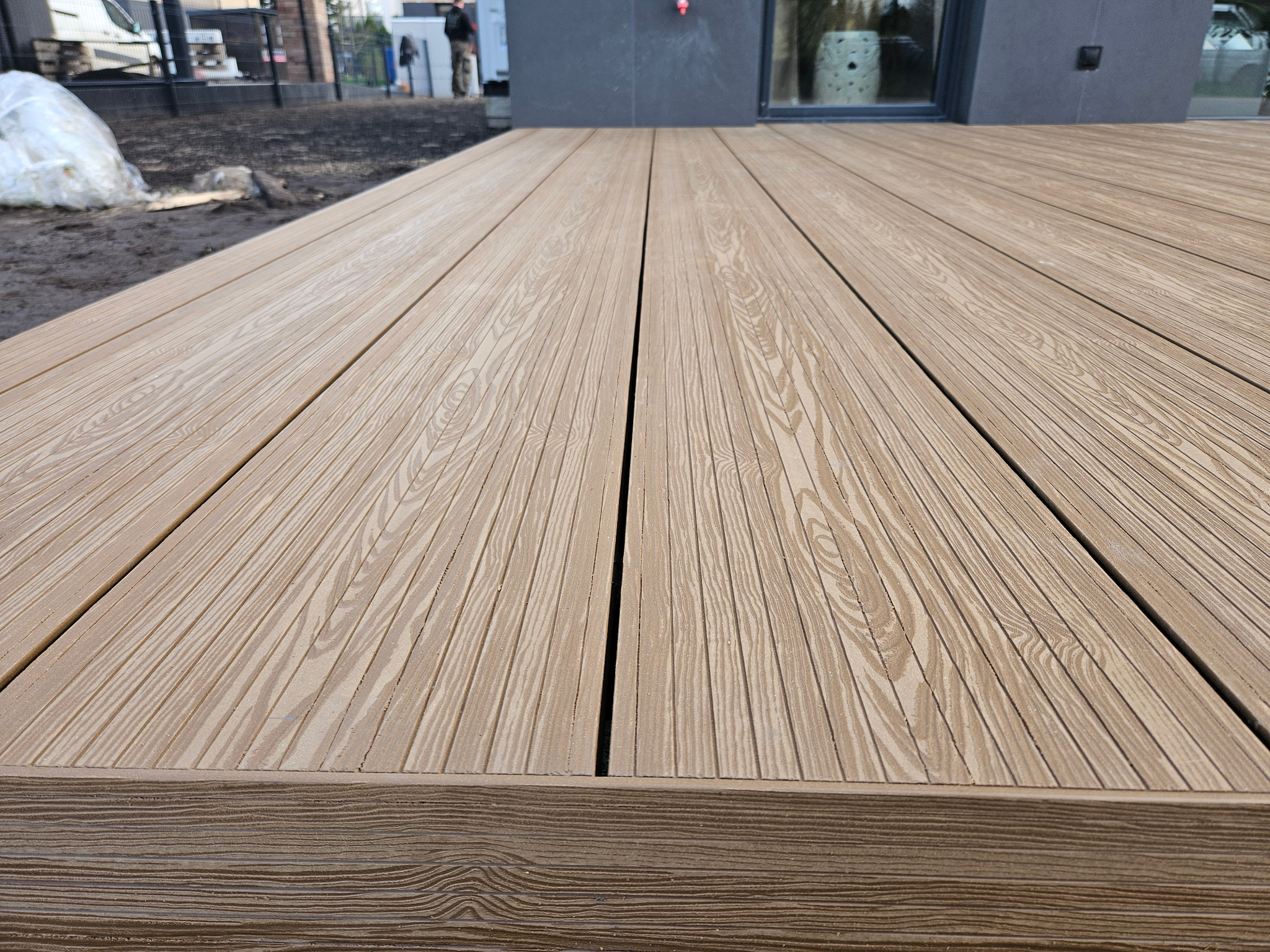 Nowo zainstalowany taras kompozytowy o jasnobrązowym odcieniu, widoczna faktura drewna, zarys budynku w tle.