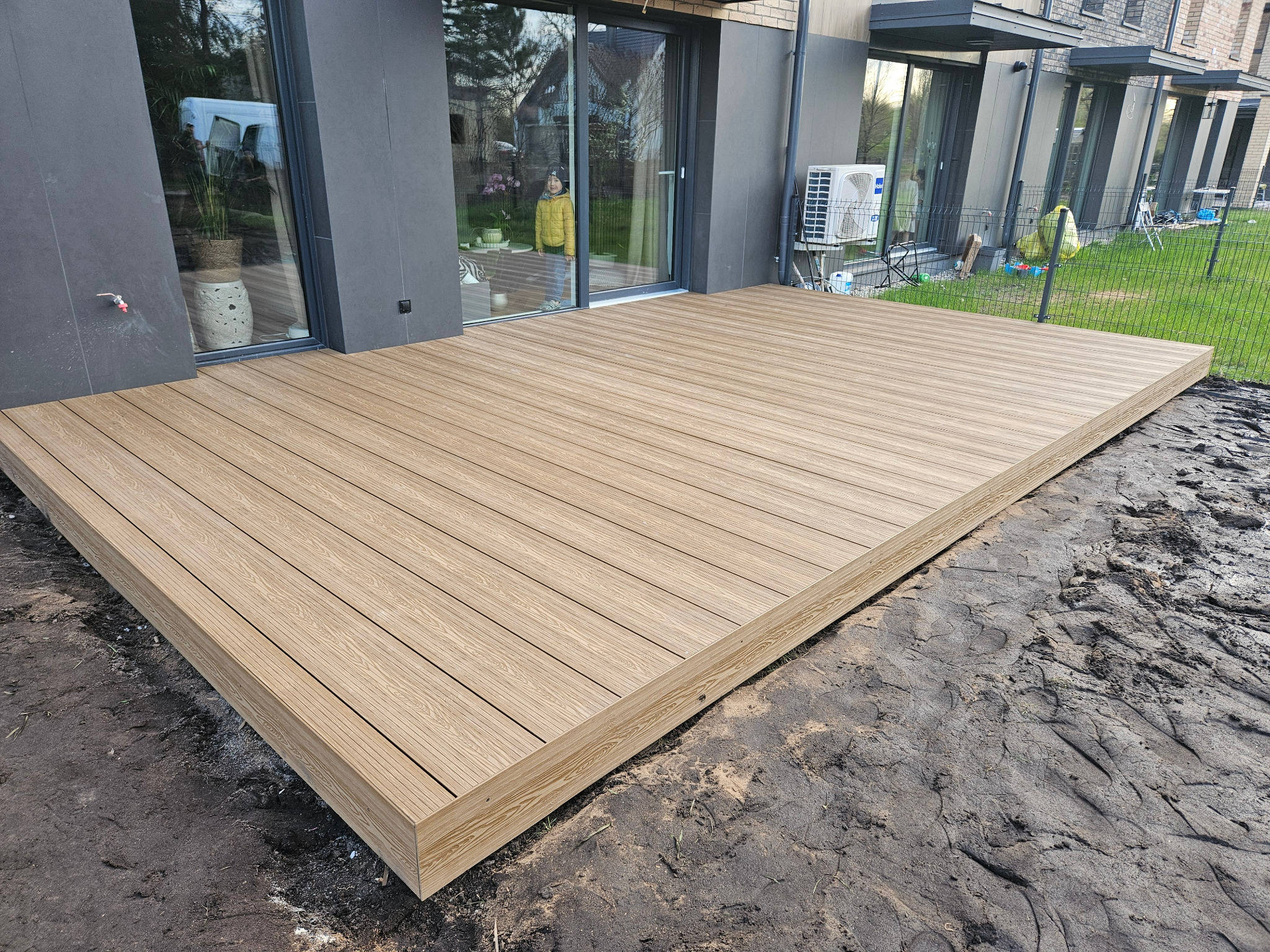 Nowoczesny taras z desek kompozytowych w odcieniach beżu, widok na szklane drzwi przesuwne i ogród w tle.