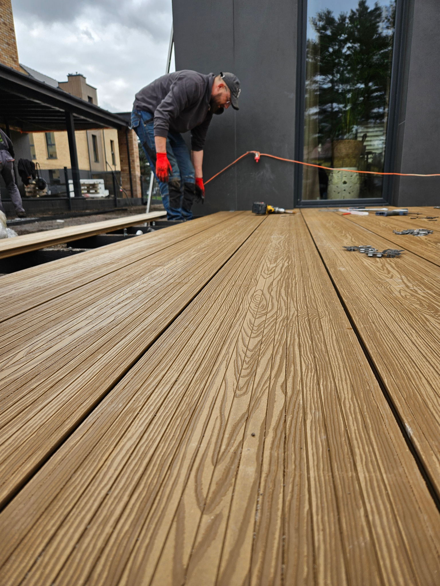 Montaż desek tarasowych: mężczyzna w czerwonych rękawicach przykuca nad nowym tarasem, widoczny wkrętak i akcesoria montażowe, tło stanowi nowoczesny budynek z dużym oknem.