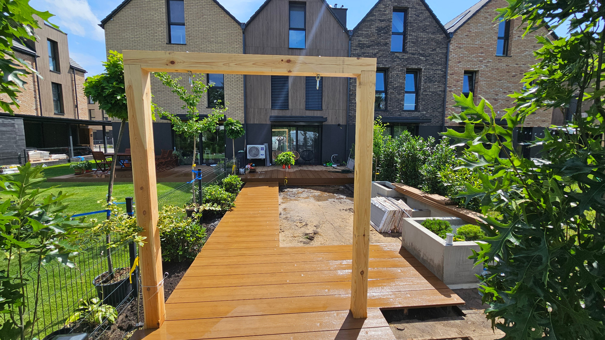 Montaż tarasu kompozytowego: widok na częściowo ukończony taras z drewnianą ramą, w tle nowoczesne domy i bujna zieleń.