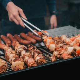 Adam Oberda - Food Group - Przygotowywanie szaszłyków i kiełbas na grillu: osoba używa szczypiec do obracania szaszłyka z kurczaka i warzyw, obok leżą przypieczone kiełbasy.